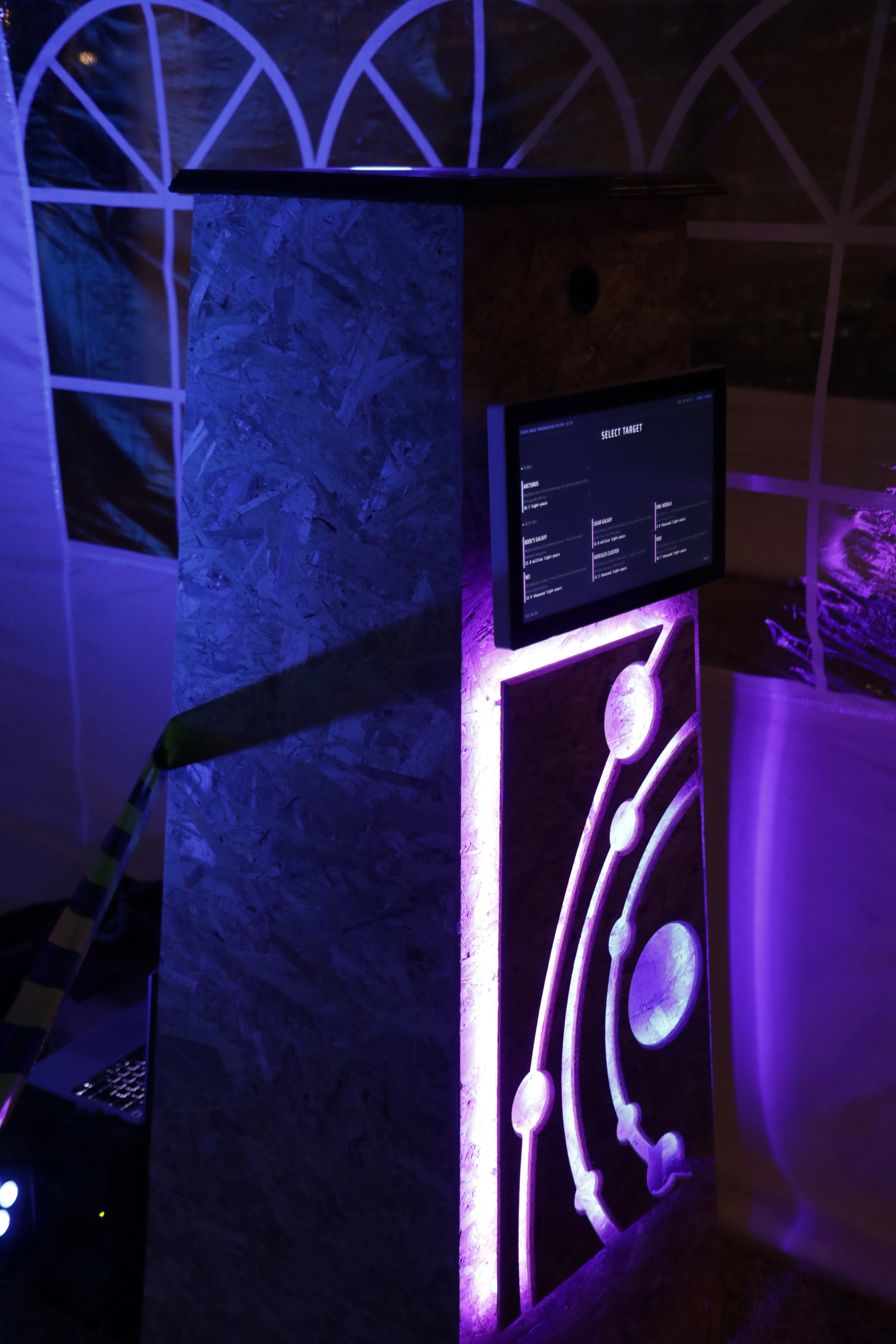 The wooden booth obelisk at night, lit from within, its touchscreen glowing against a purple-lit tent wall.